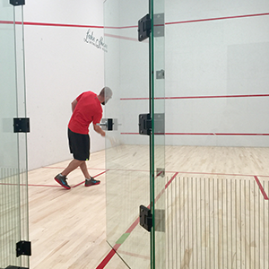 Solo squash player swinging at ball in glass paneled court.