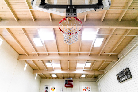View of basketball gym ceiling and basketball hoop. Light shining in through skylights.